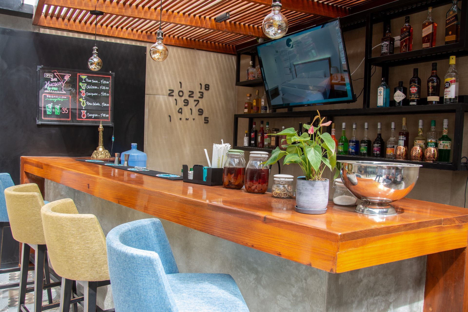 A modern bar with a wooden counter, blue and yellow stools, a television screen, and shelves filled with liquor bottles.