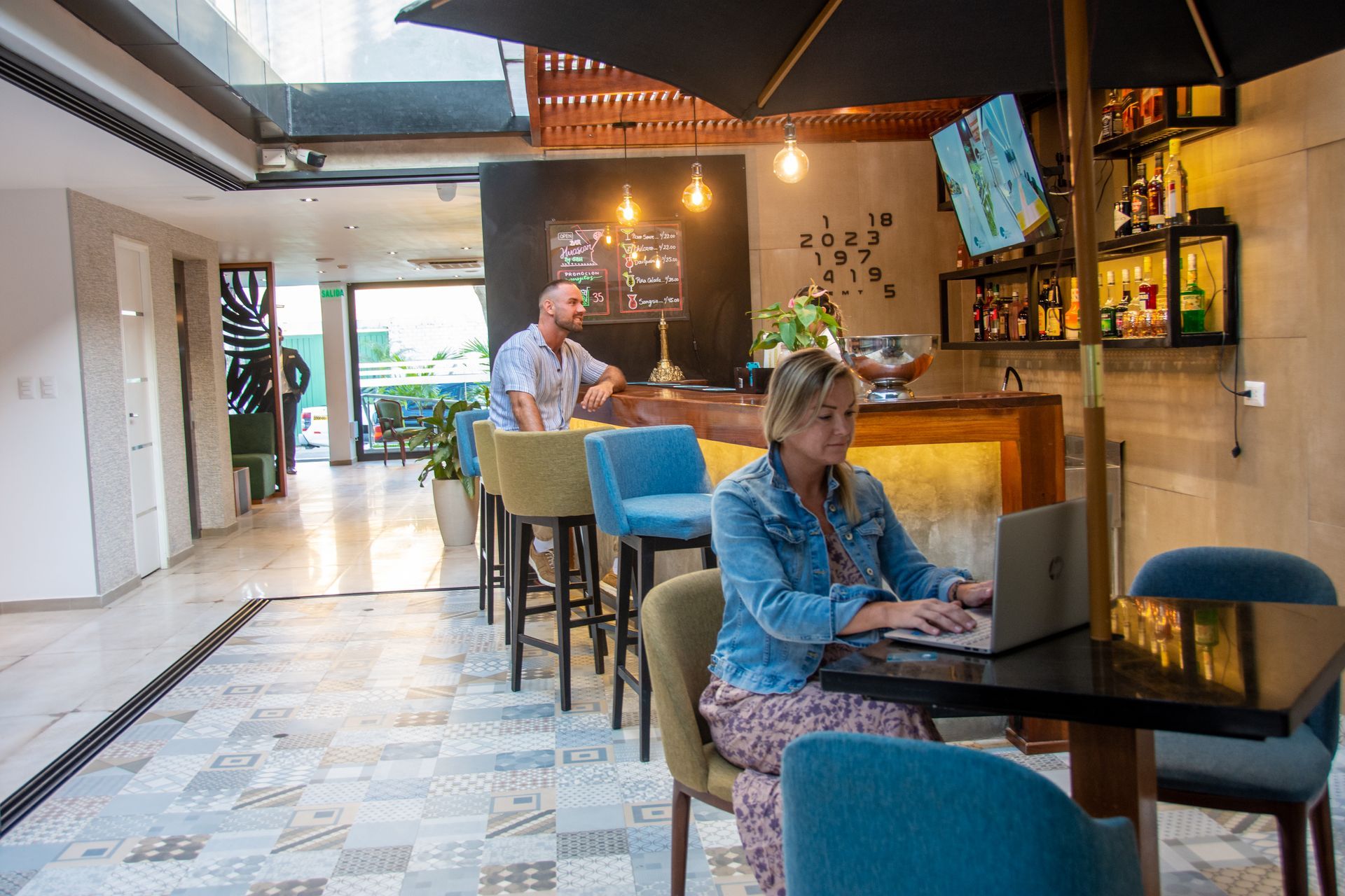 A woman uses a laptop at a small table in a bar area, while a person sits at the counter in the background.