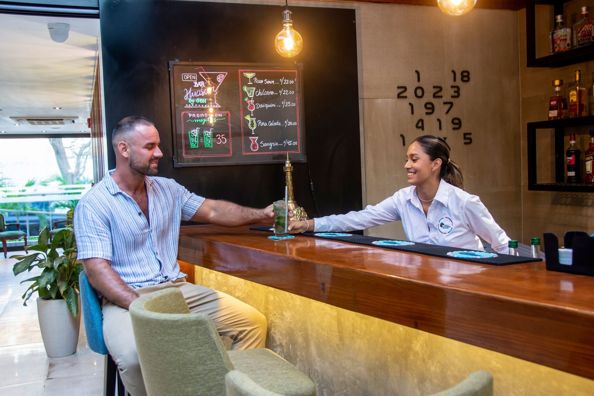 A bartender serves a cocktail to a patron sitting at a well-lit bar counter.