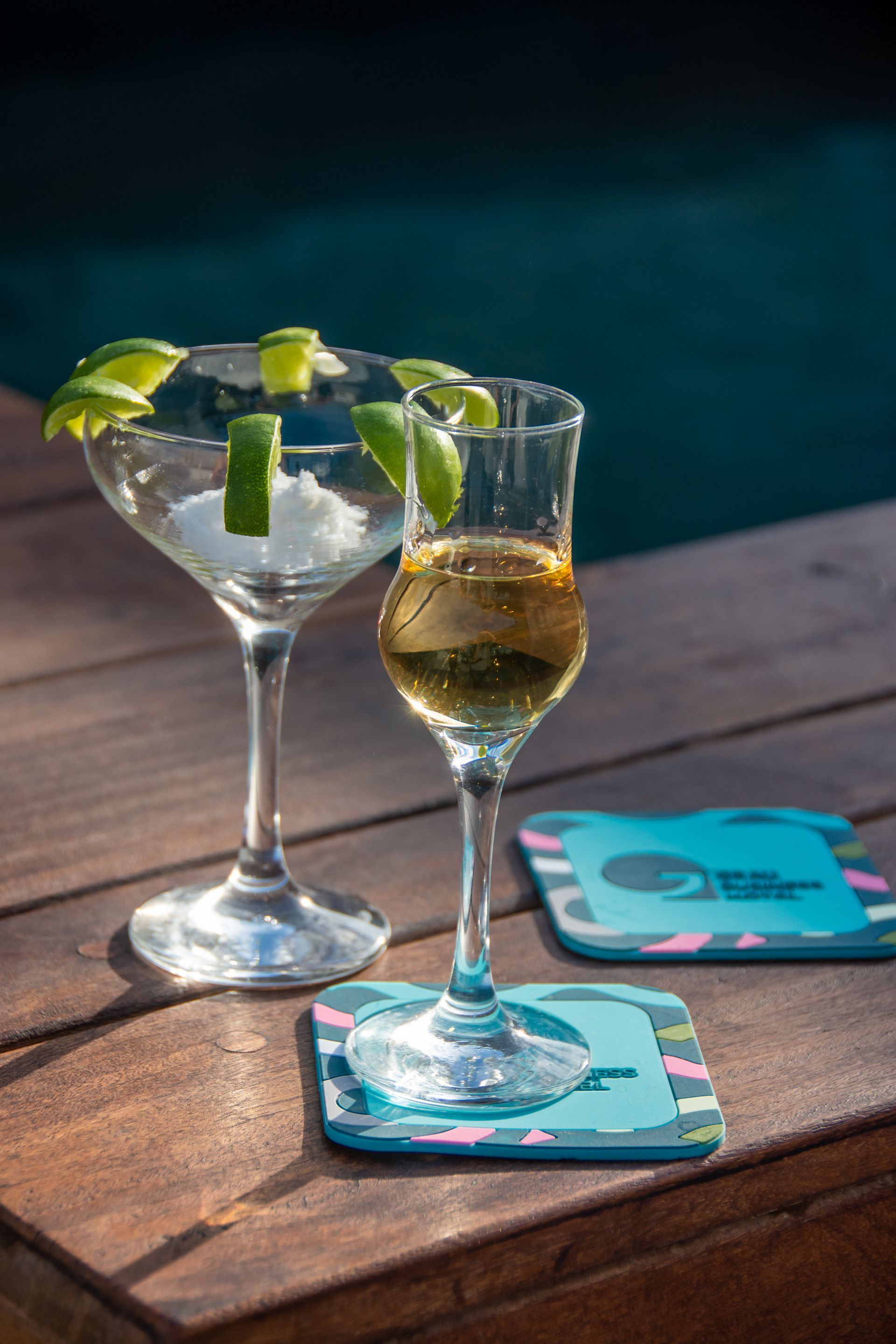 A martini glass garnished with lime, a filled shot glass, and patterned coasters sit on a wooden deck by a pool.