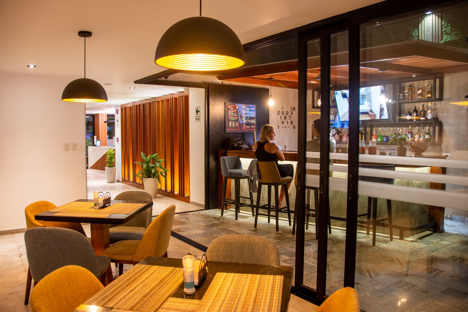 A restaurant interior with dining tables in the foreground and a bar area with stools in the background under warm lights.