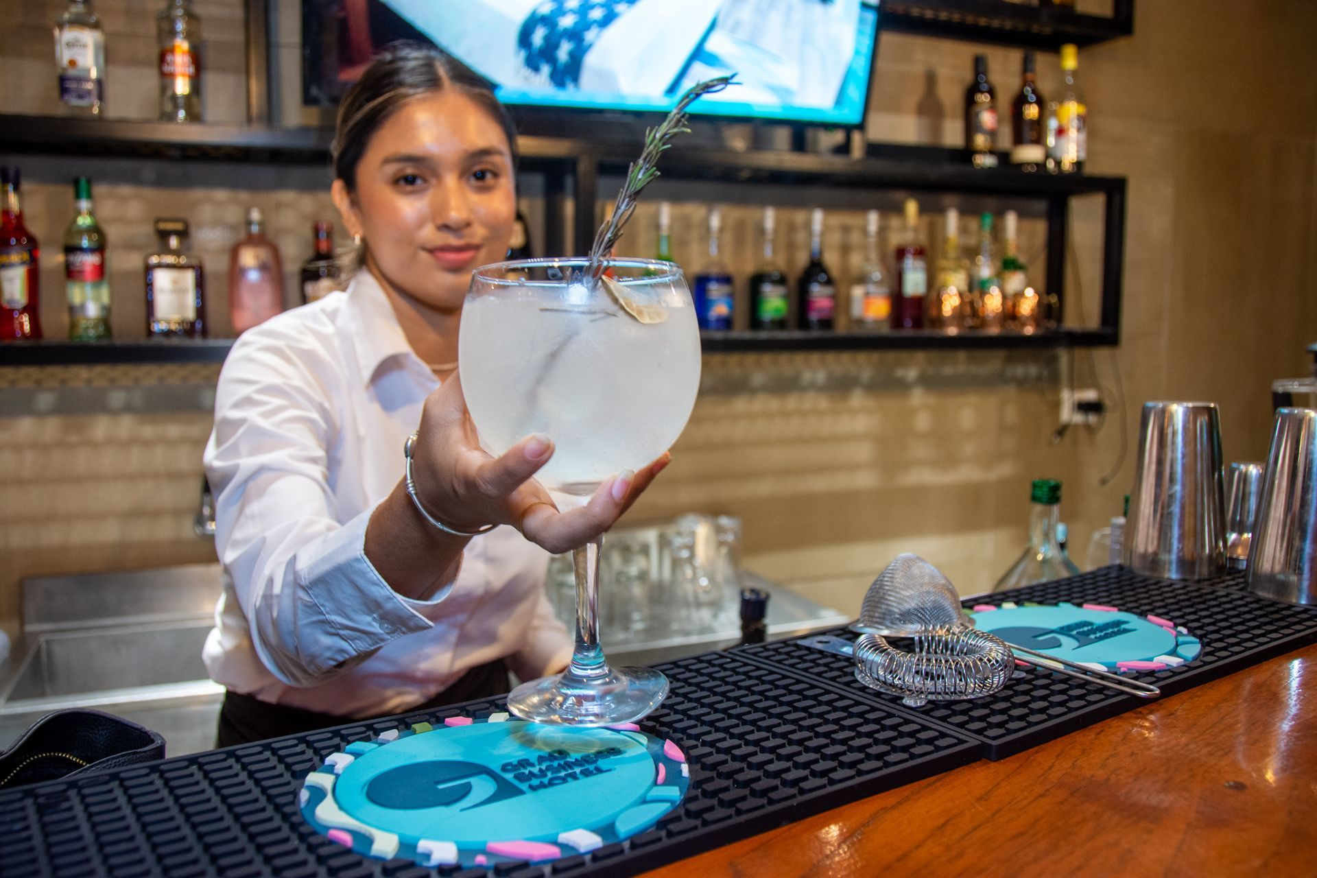 A bartender holding up a large stemmed glass filled with a clear cocktail and a garnish, at a bar with bottle shelves.