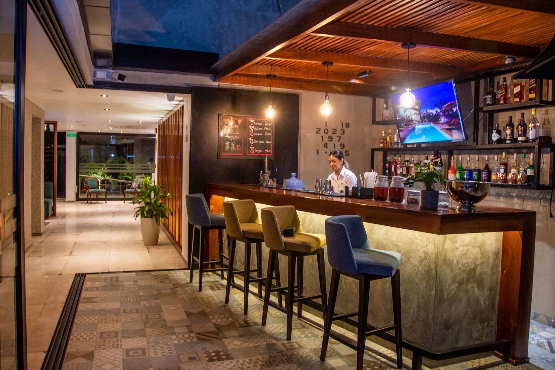 A modern bar interior with a bartender, cushioned stools at a wooden counter, decorative lighting, and patterned flooring.