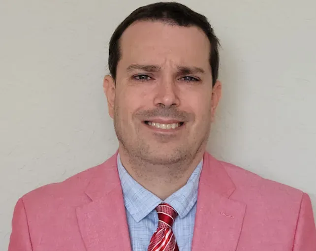 Man in pink suit, blue shirt, and patterned red tie smiling in front of a neutral background.