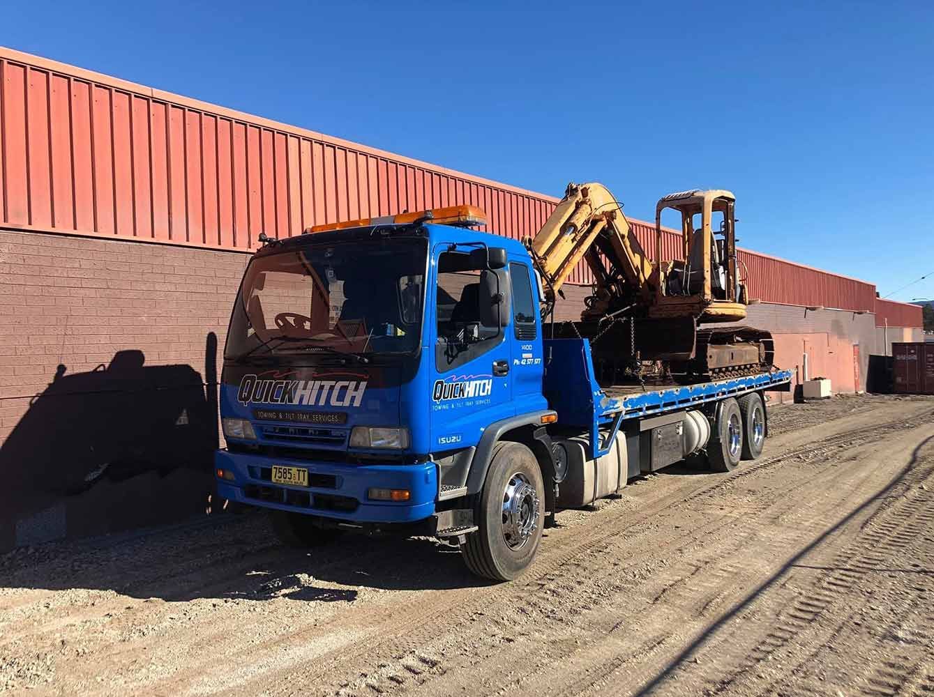 A Blue Tow Truck is Towing a Yellow Excavator — Quick Hitch Towing & Tilt Tray Services Illawarra in Oak Flats, NSW