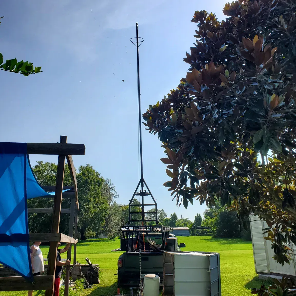 Tall metal mast in a grassy yard beside trees and equipment under a clear blue sky