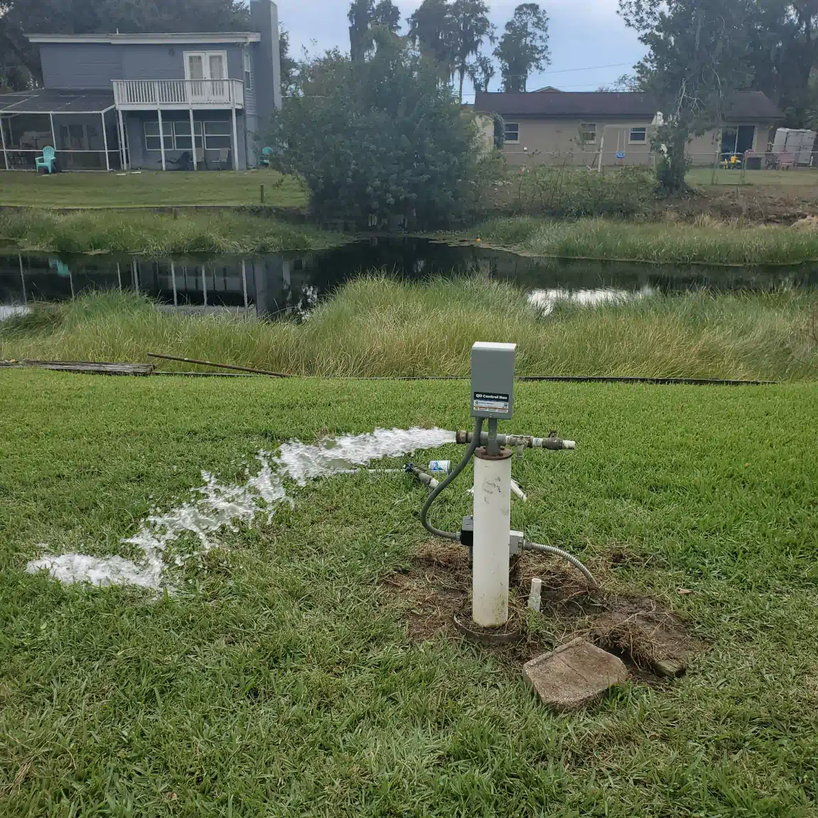 Sprinkler spraying water across a grassy yard beside a pond and houses