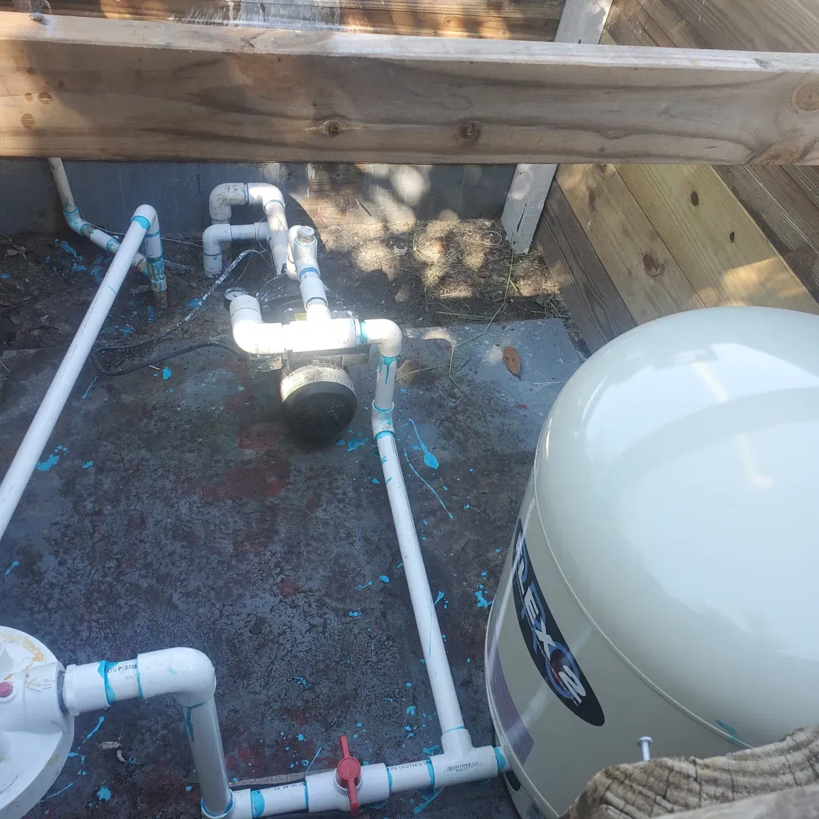 Backyard pool equipment with white PVC pipes, valves, and a beige filter tank under a wooden deck.