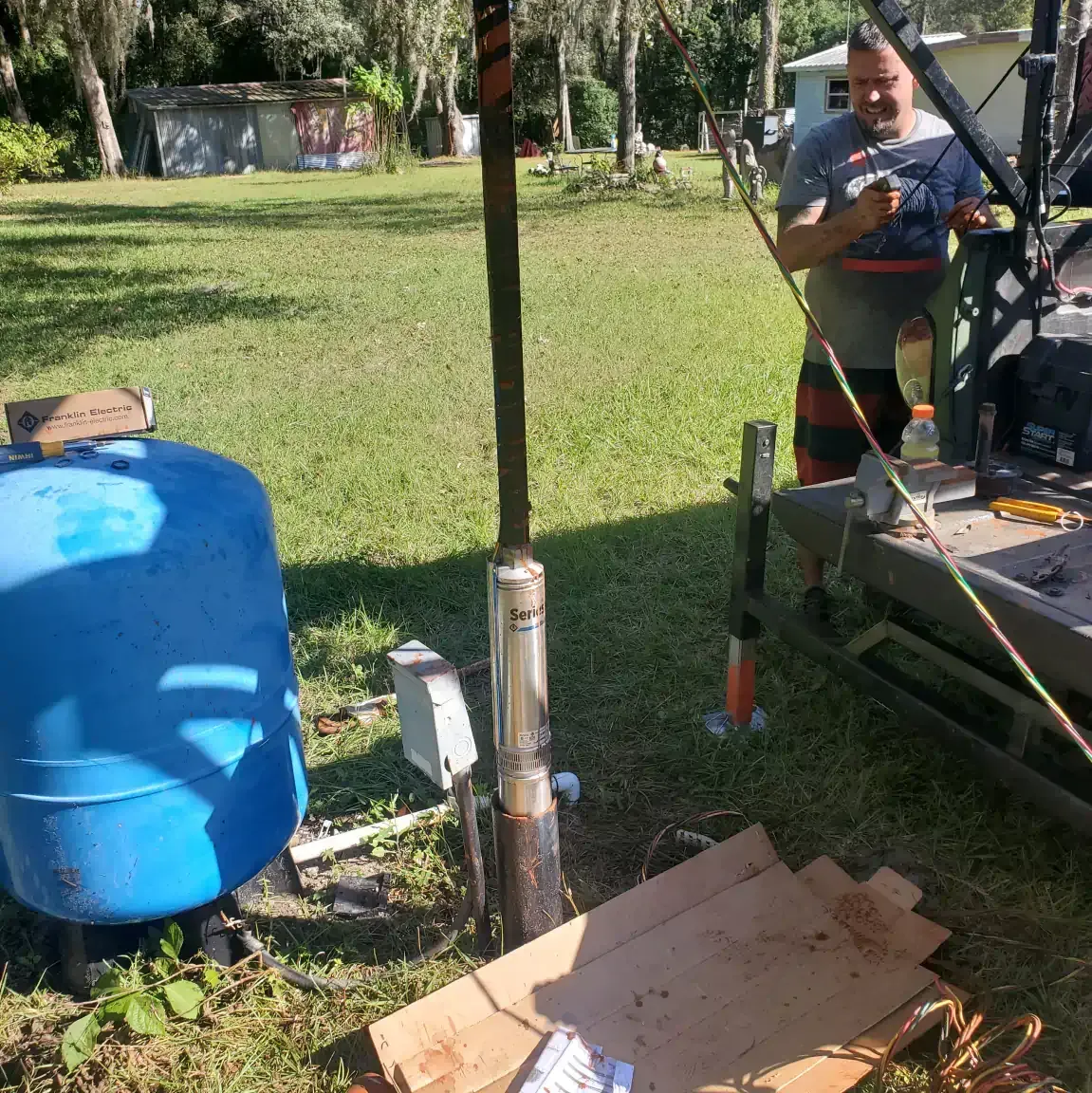 Person operating machinery outdoors beside a blue barrel and equipment on a grassy lawn