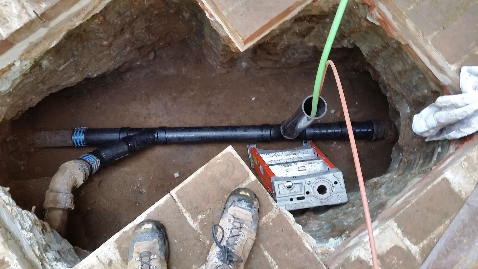 Plumbing repair in a brick-lined pit: black pipes, tan elbow joint, tools, and a person's muddy boots.