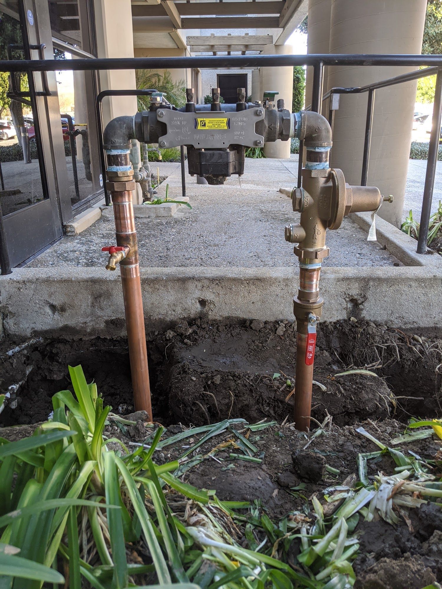 Two upright copper pipes with valves and a backflow preventer in a dirt bed next to a building.