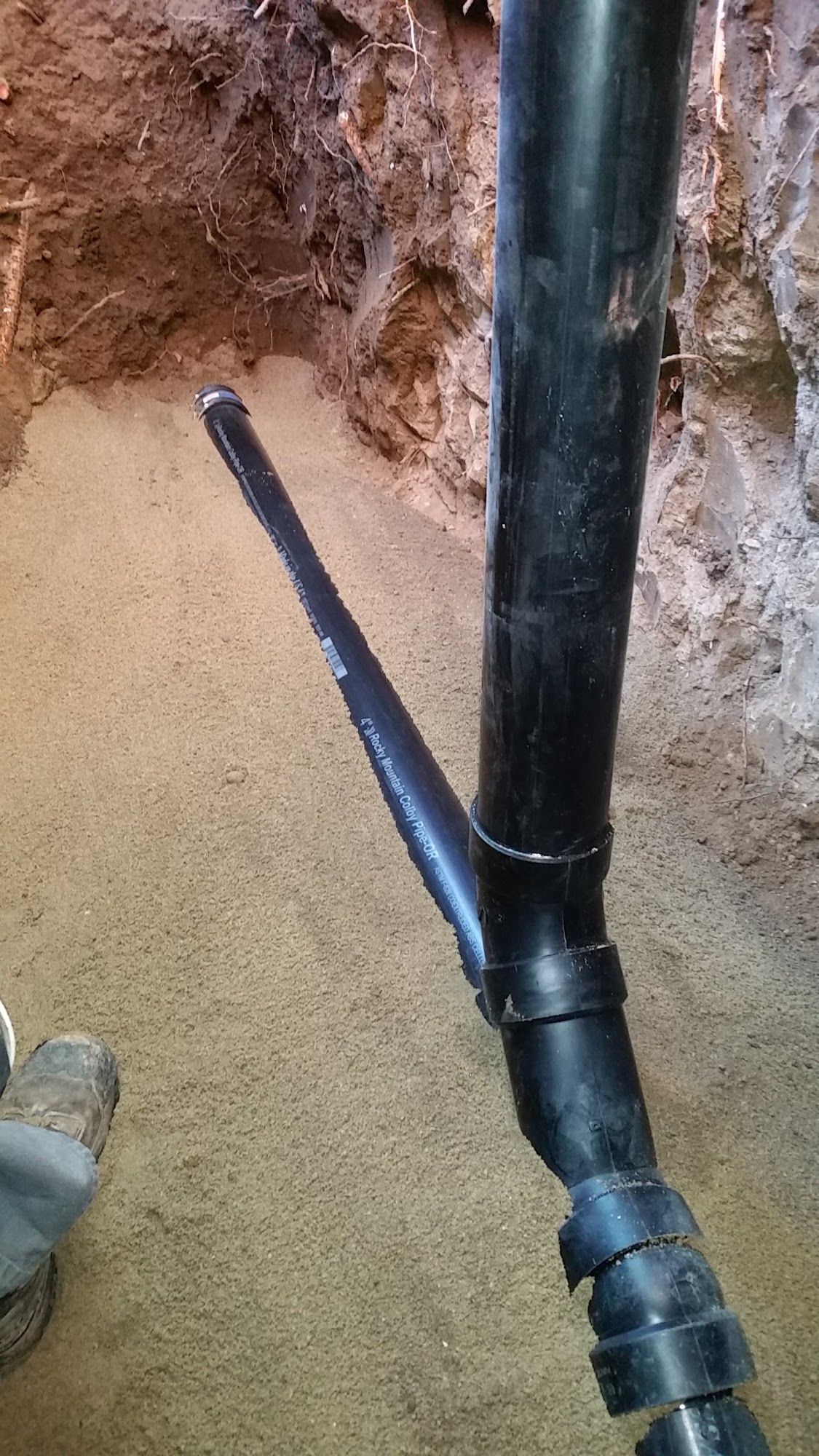 Black plumbing pipes in a trench, connected with angled joints, against dirt and sand.