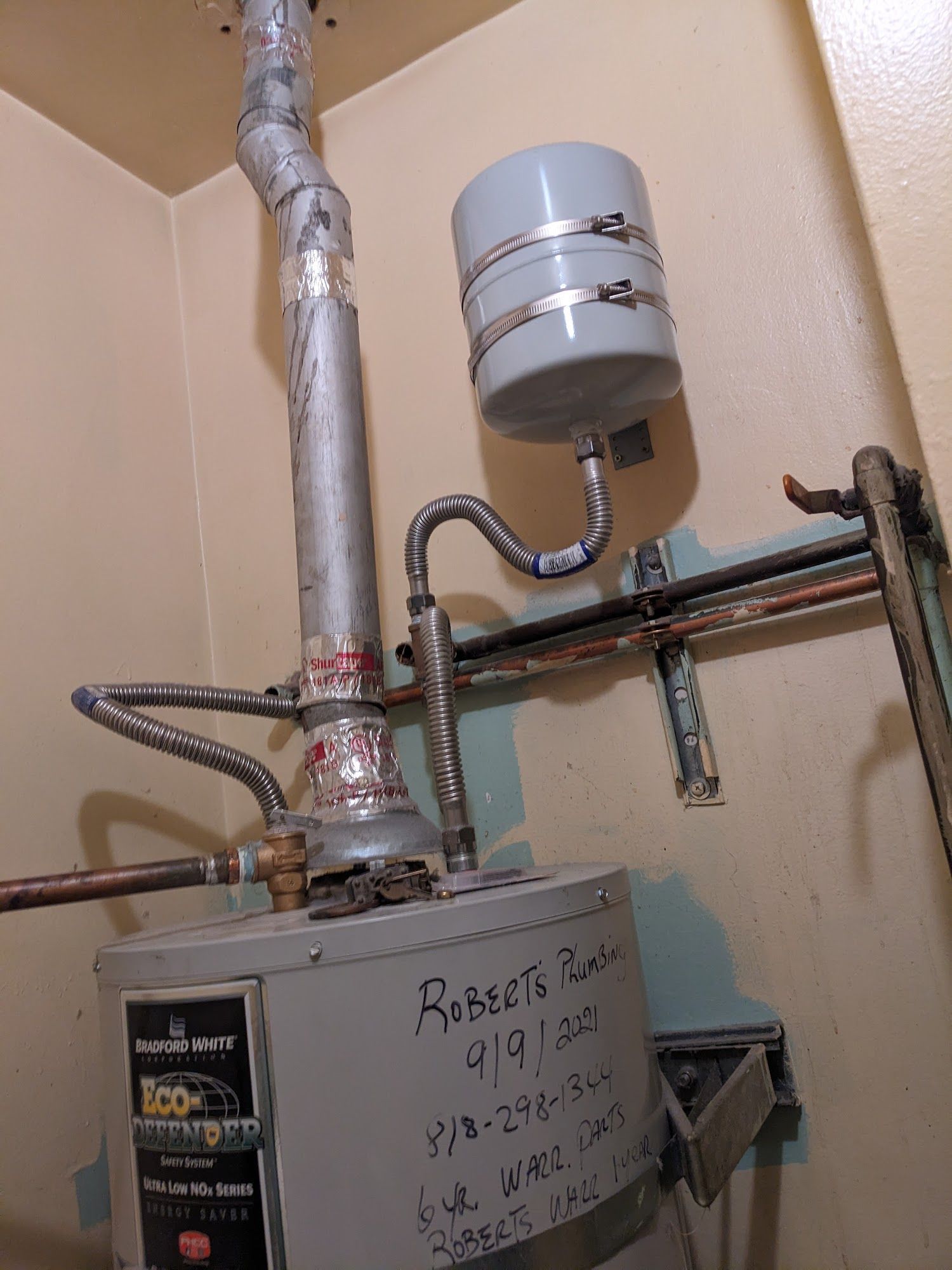 A water heater with a connected expansion tank and piping, mounted against a beige wall.