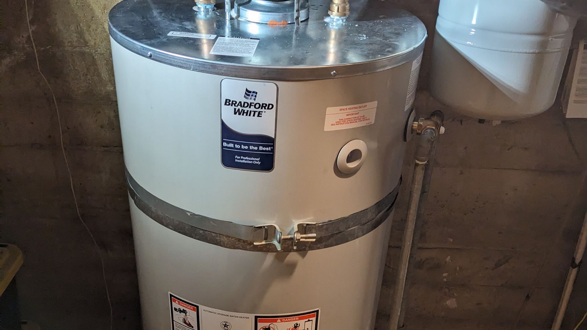 Water heater in a basement. Cylindrical, silver top, white body with blue label. Water pipes connected.