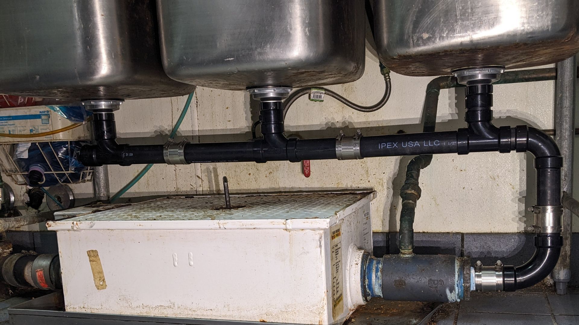 Underside of three stainless steel sinks with black plumbing connected to a white grease trap in a commercial kitchen.