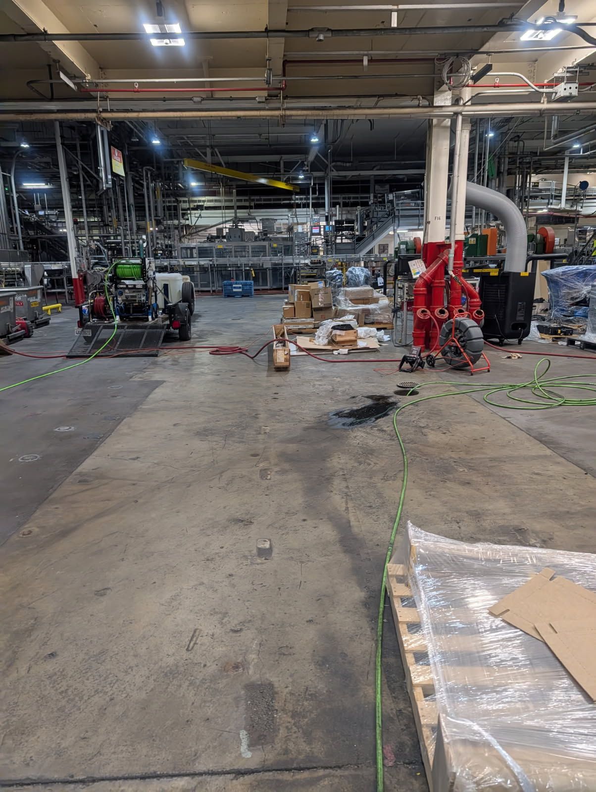 Cluttered warehouse floor with machinery, pallets, and ducting under overhead lights