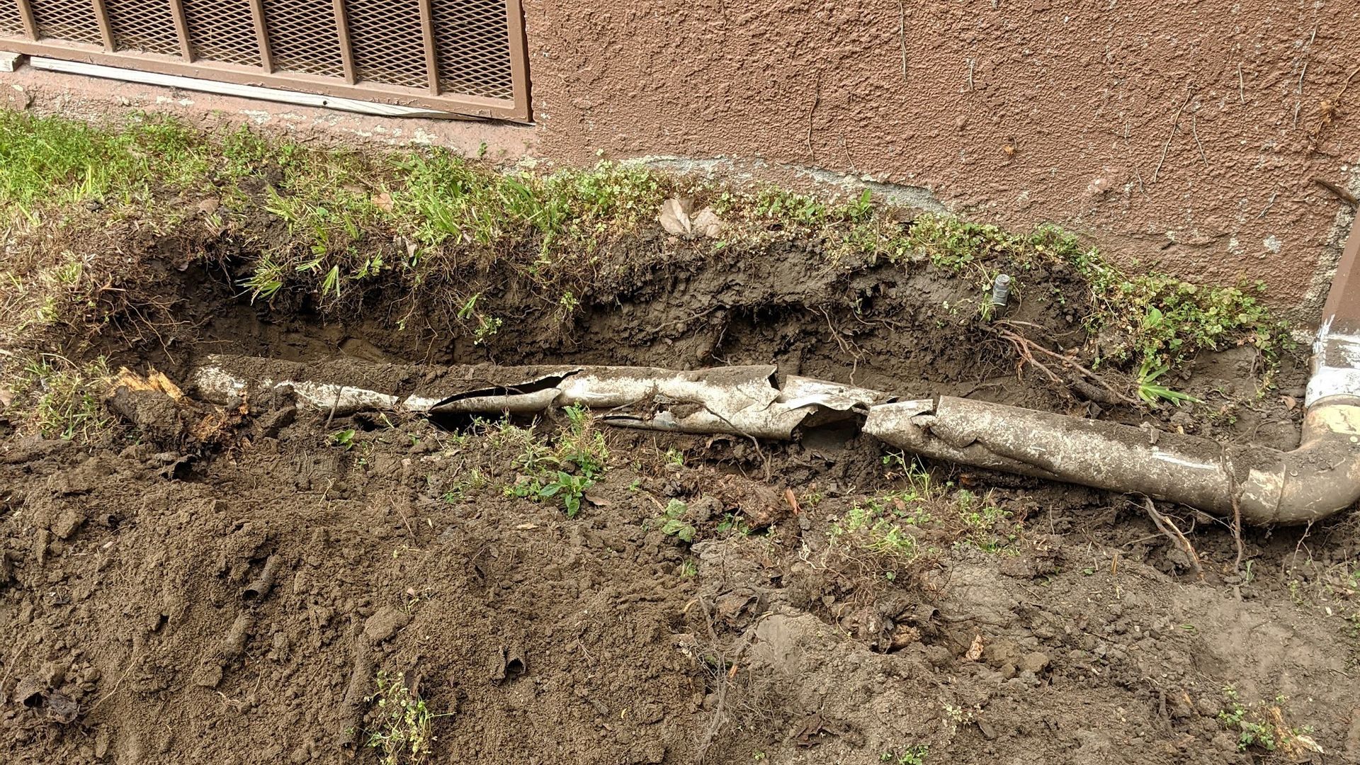 Damaged pipe exposed in a trench near a building.