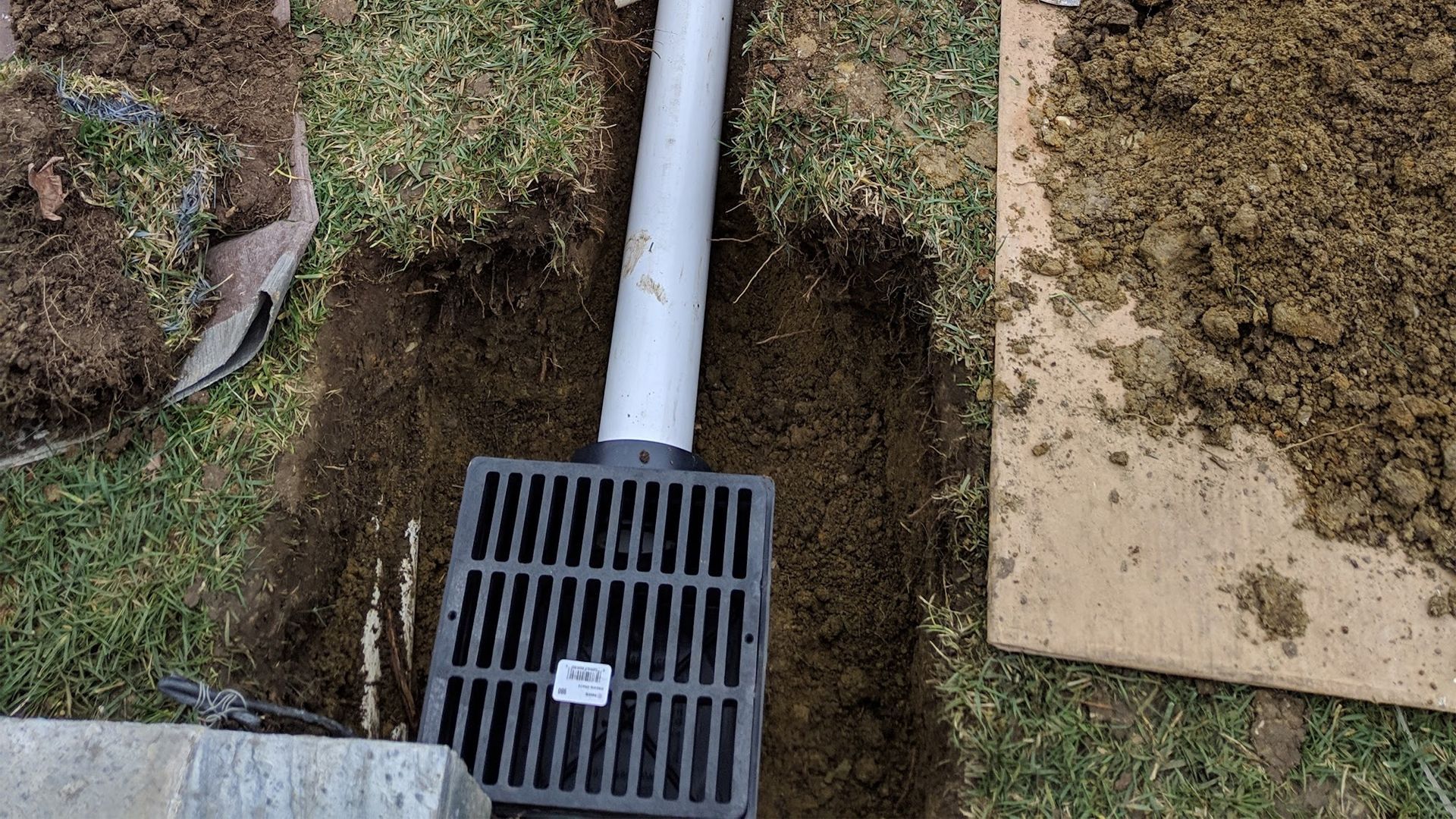 Drainage grate with connecting pipe in a dirt trench, surrounded by grass and soil.