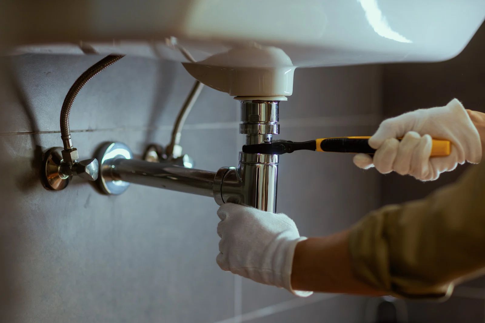 Person wearing gloves working on bathroom sink plumbing with a tool.
