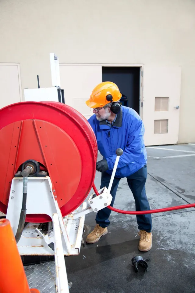 Man in a blue jacket and hard hat operates a red hose reel outdoors, looking intently at the equipment.