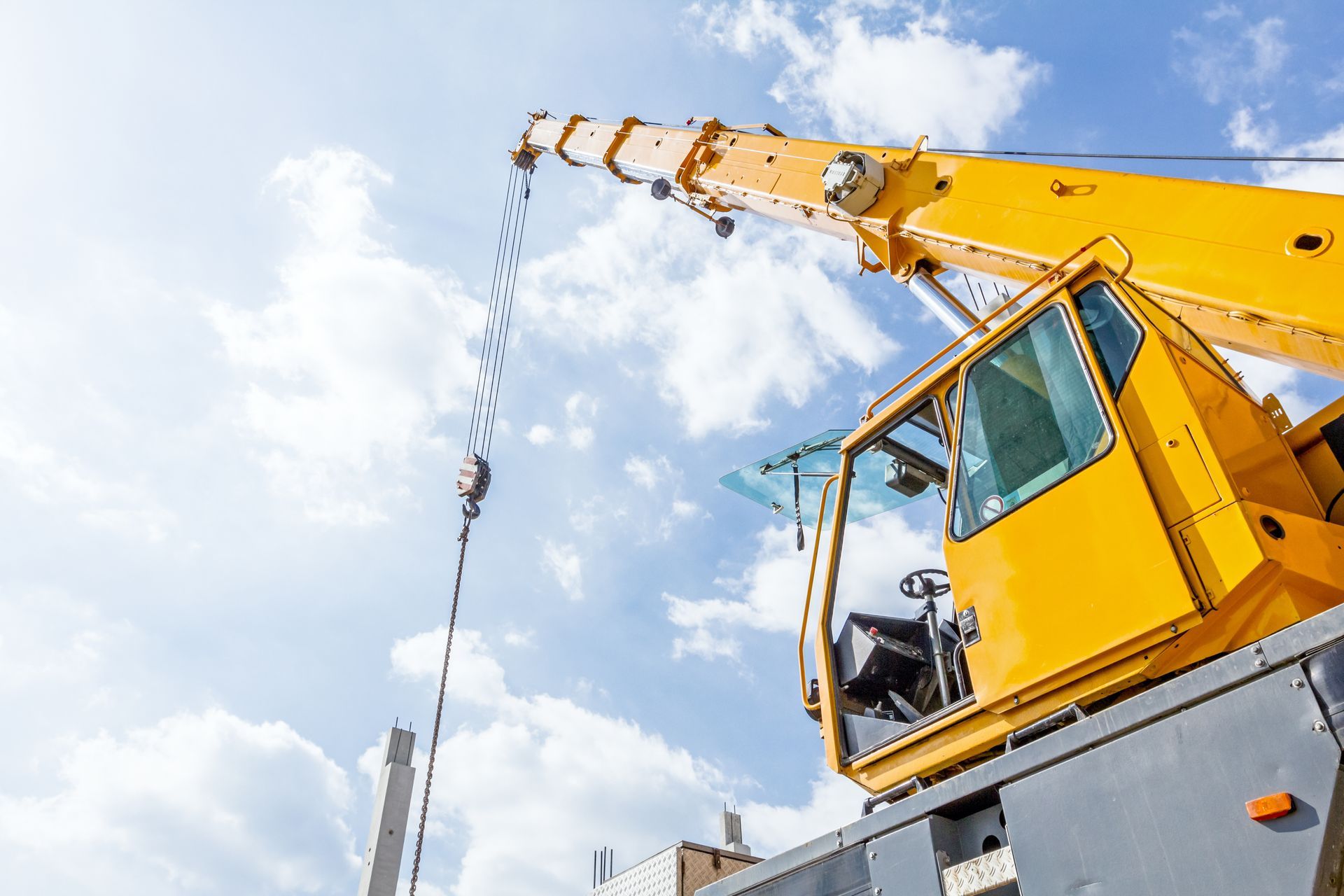 Yellow crane against a cloudy blue sky, lifting with a suspended hook over a construction site.