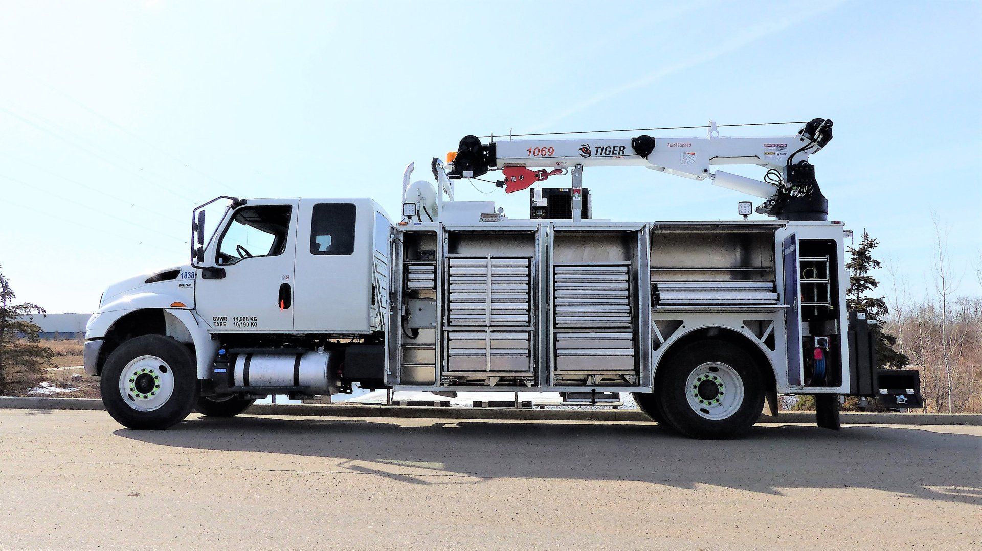 A white utility truck with a mounted crane and storage compartments parked outside against a clear blue sky.
