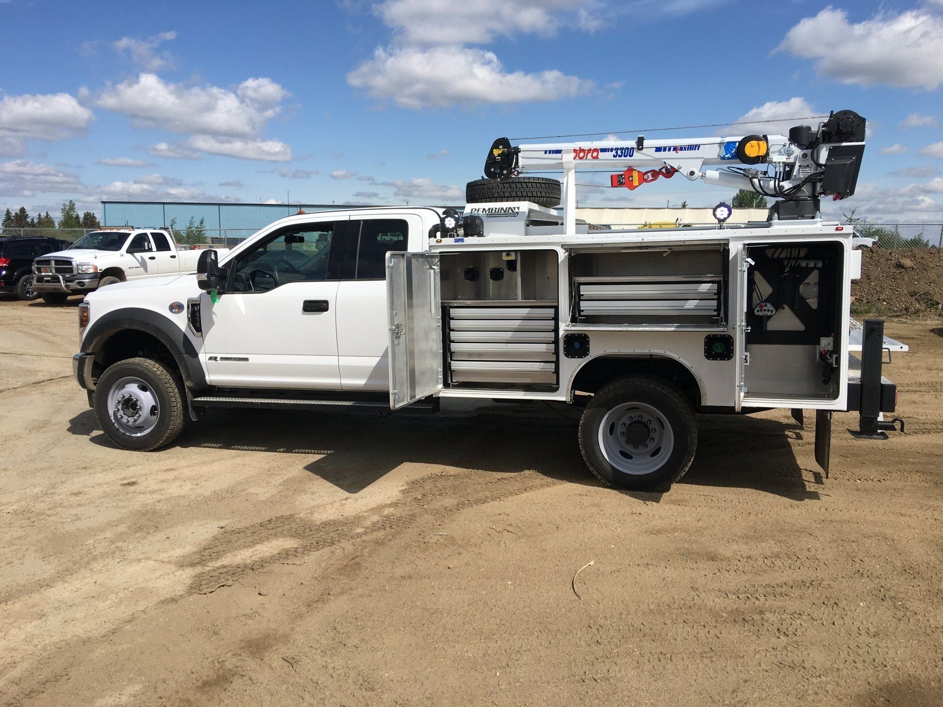White work truck with open compartments, crane, and tools on a dirt lot.