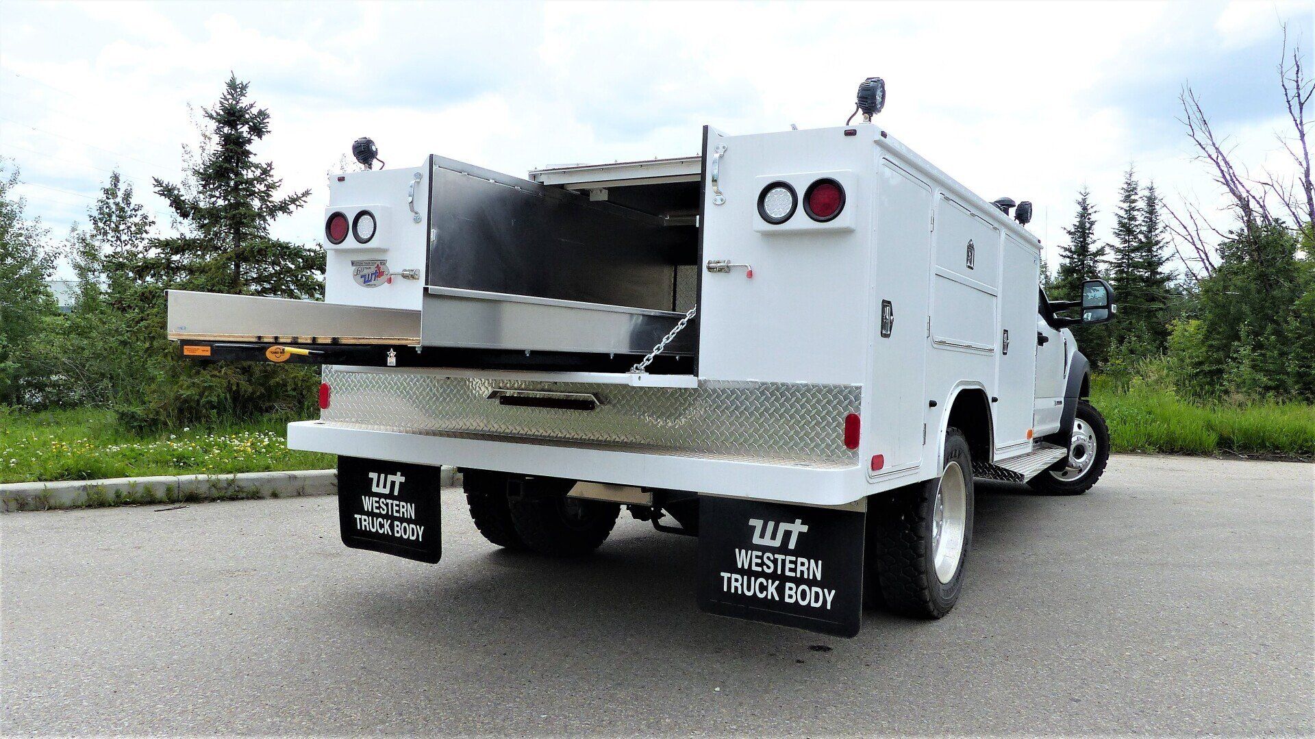A white utility service truck with an extended rear cargo slide, parked on asphalt with trees in the background.