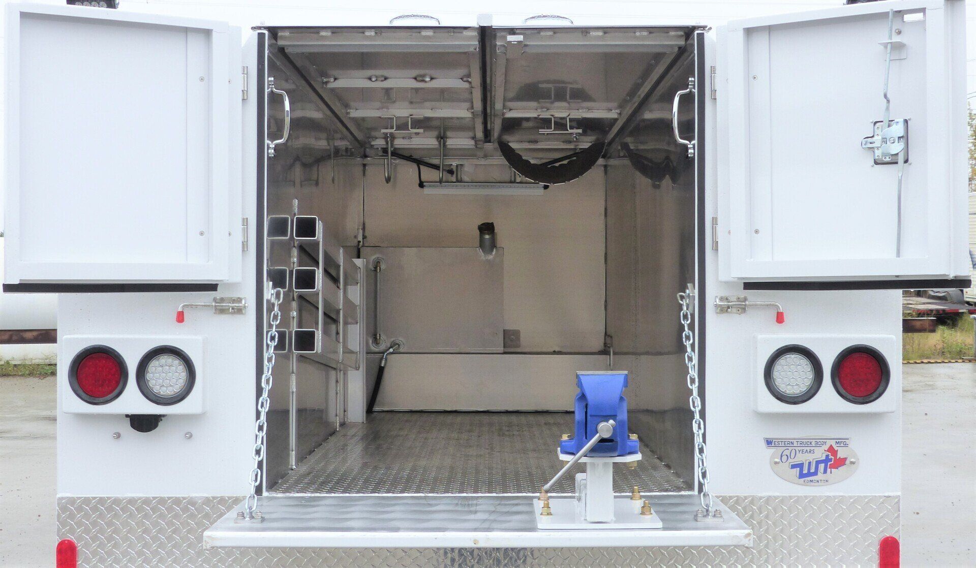 The rear view of a white utility truck with open doors, showing an empty cargo bay, interior shelving, and a blue vise.