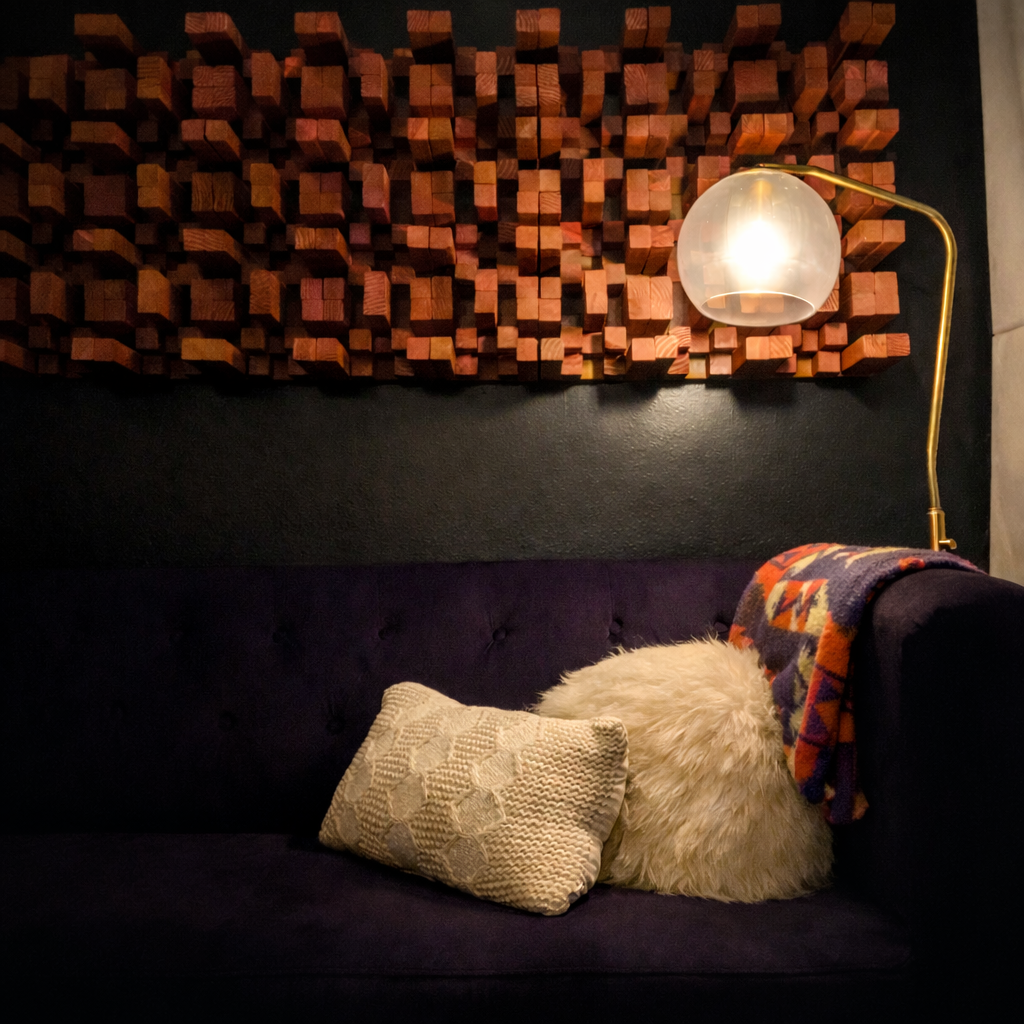 A dark purple sofa with two throw pillows and a patterned blanket, set beneath a textured wooden wall art and a gold lamp.