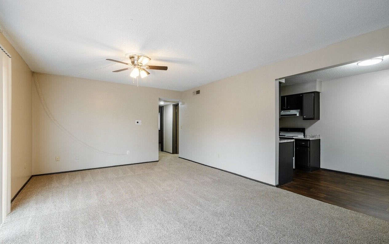 Empty living room with beige walls and carpet, adjacent to a kitchen area. Ceiling fan visible.