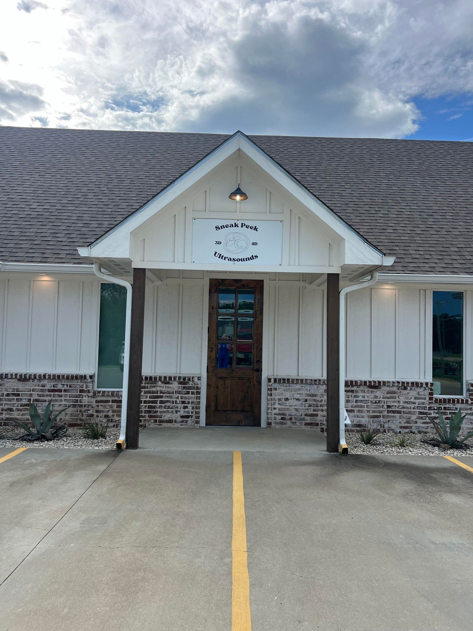 Front view of a small white building with a central door, shingled roof, and paved entry under a cloudy sky