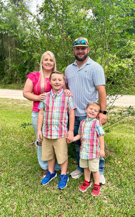Family of four standing outdoors on grass, smiling in colorful summer clothes.