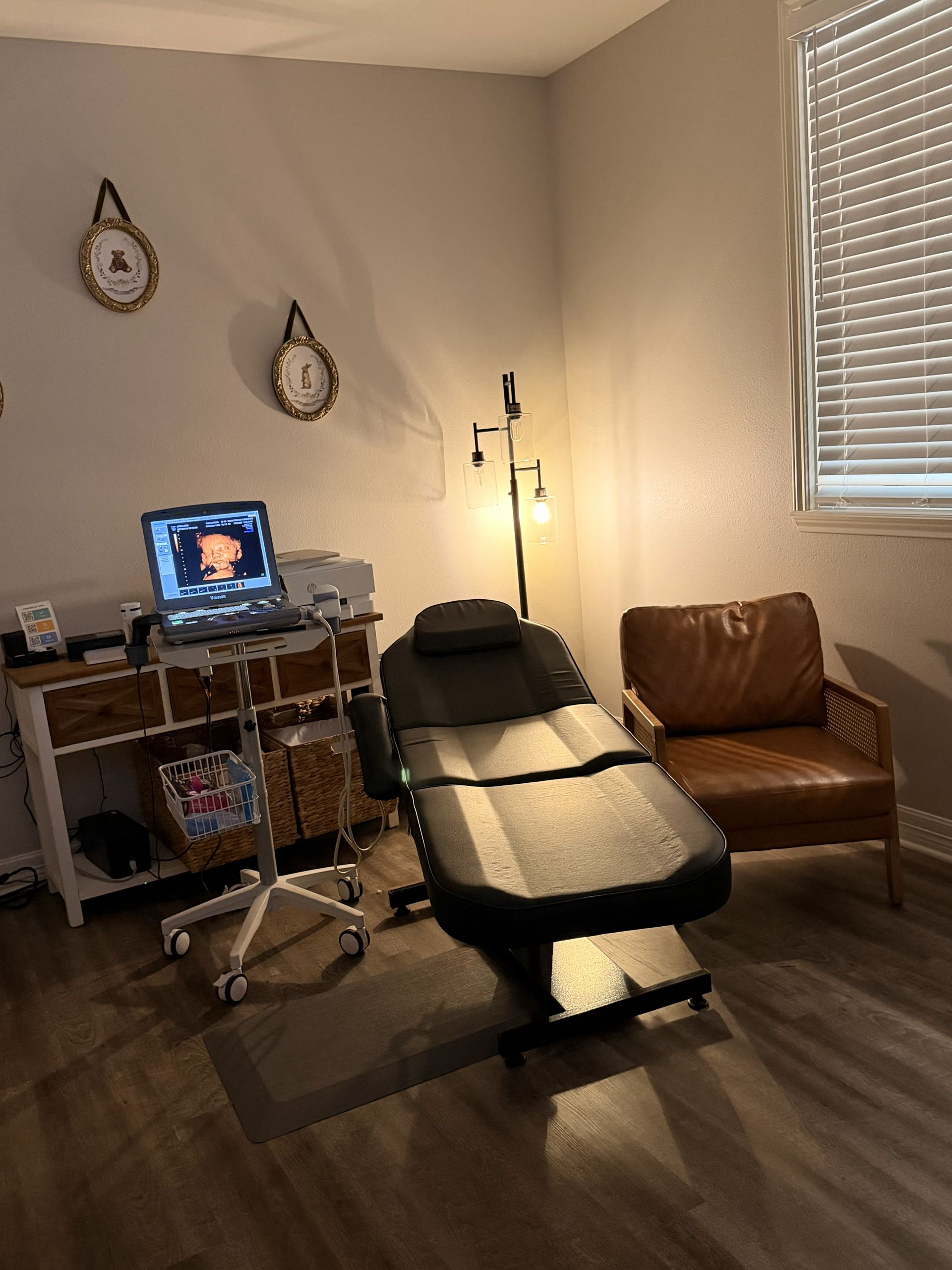 office with ultrasound bed, desk, laptop, floor lamp, and brown chair by a window.