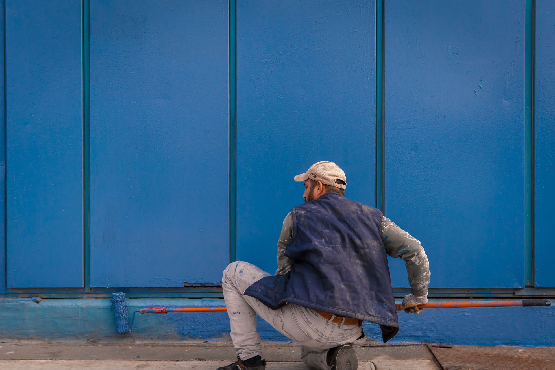 Worker crouching beside a large blue wall, wearing a cap, jacket, and light pants.
