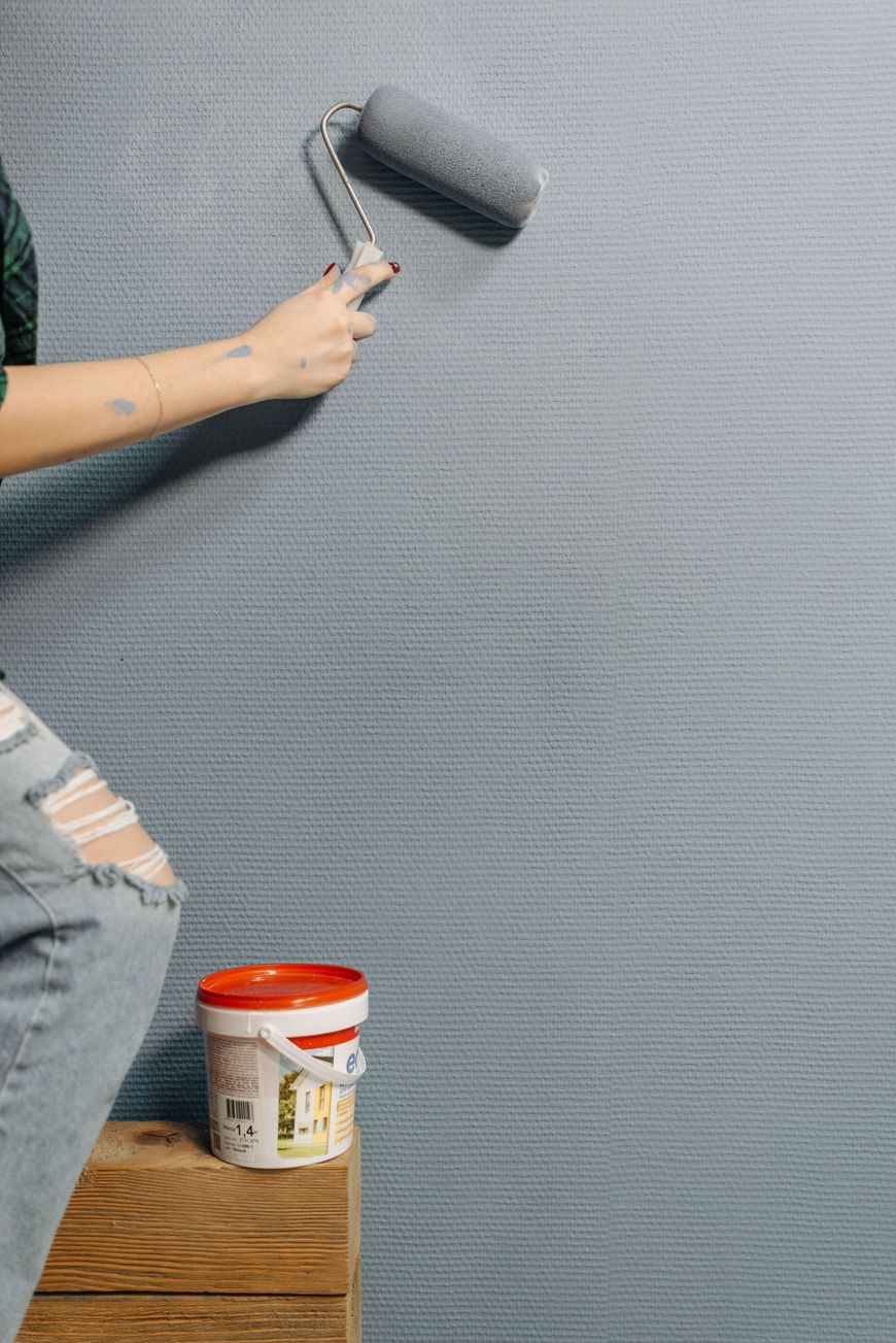 Person painting a gray wall with a roller beside a paint can on a wooden floor.
