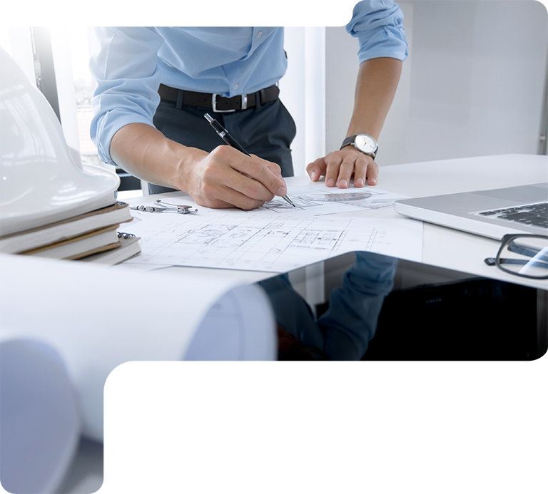Person in a blue shirt sketching plans on a desk with a laptop and folded blueprints nearby