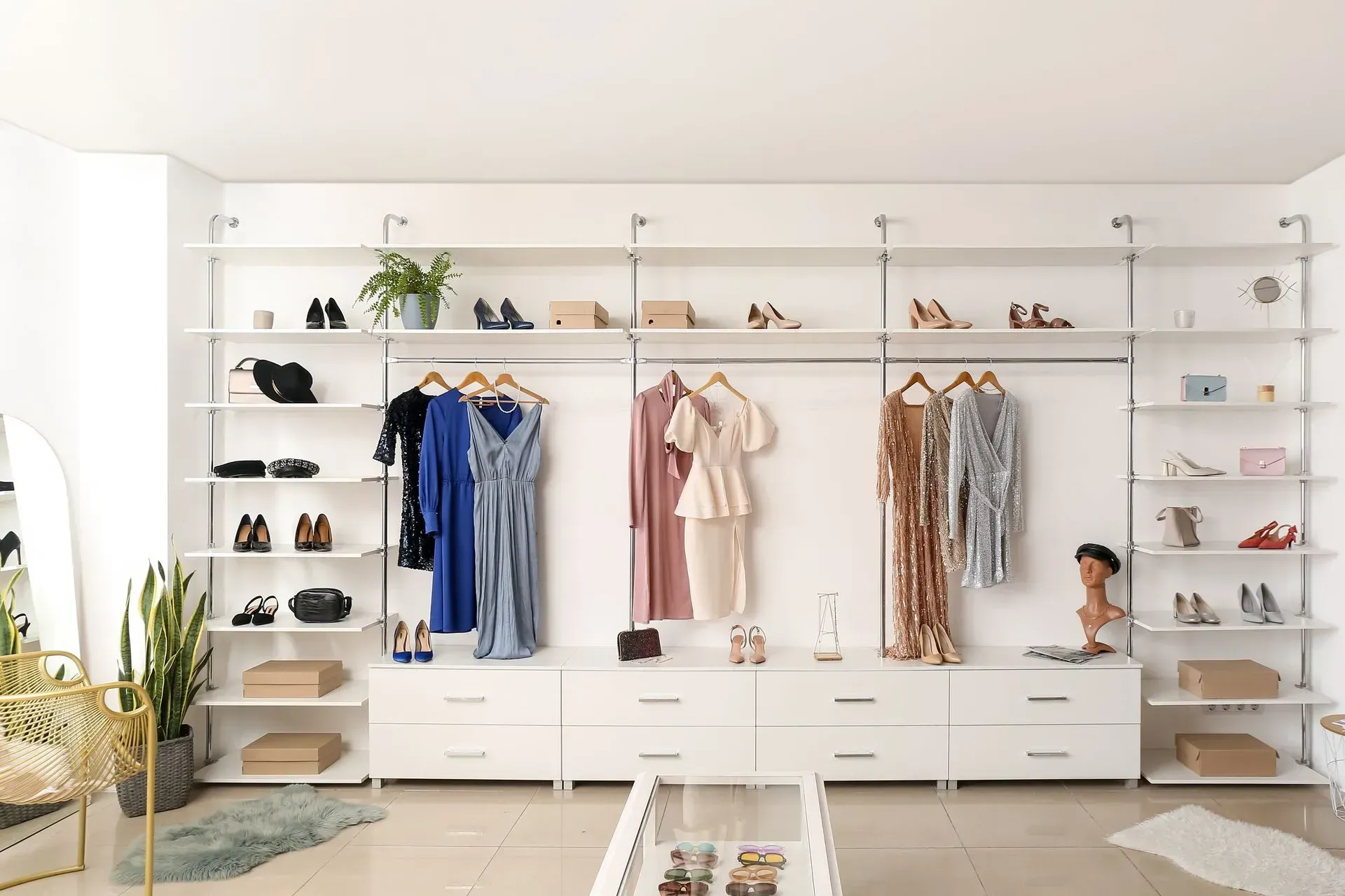 Minimalist walk-in closet with white shelves, hanging clothes, shoes, plants, and a small doll on the right.