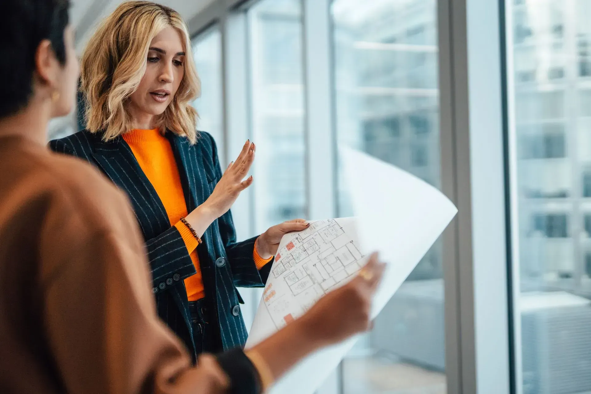 Woman in orange sweater presenting documents during office discussion beside a glass wall