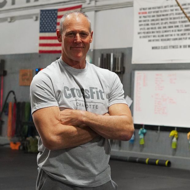 A man wearing a crossfit shirt is standing in a gym with his arms crossed