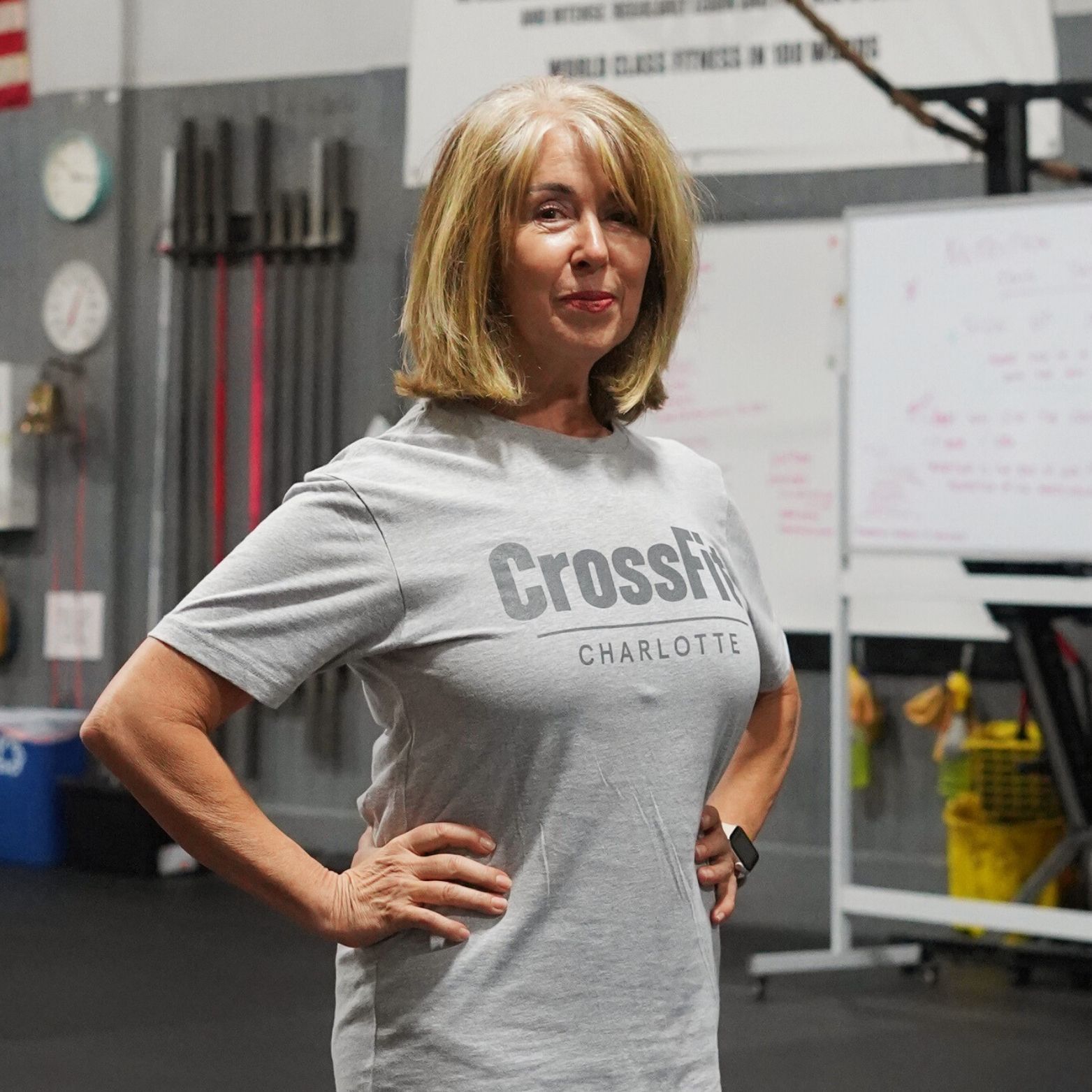 A woman wearing a grey crossfit shirt is standing in a gym with her hands on her hips.
