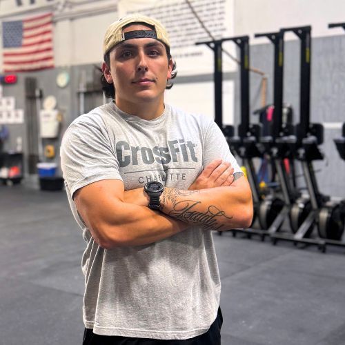 A man wearing a crossfit shirt is standing in a gym with his arms crossed