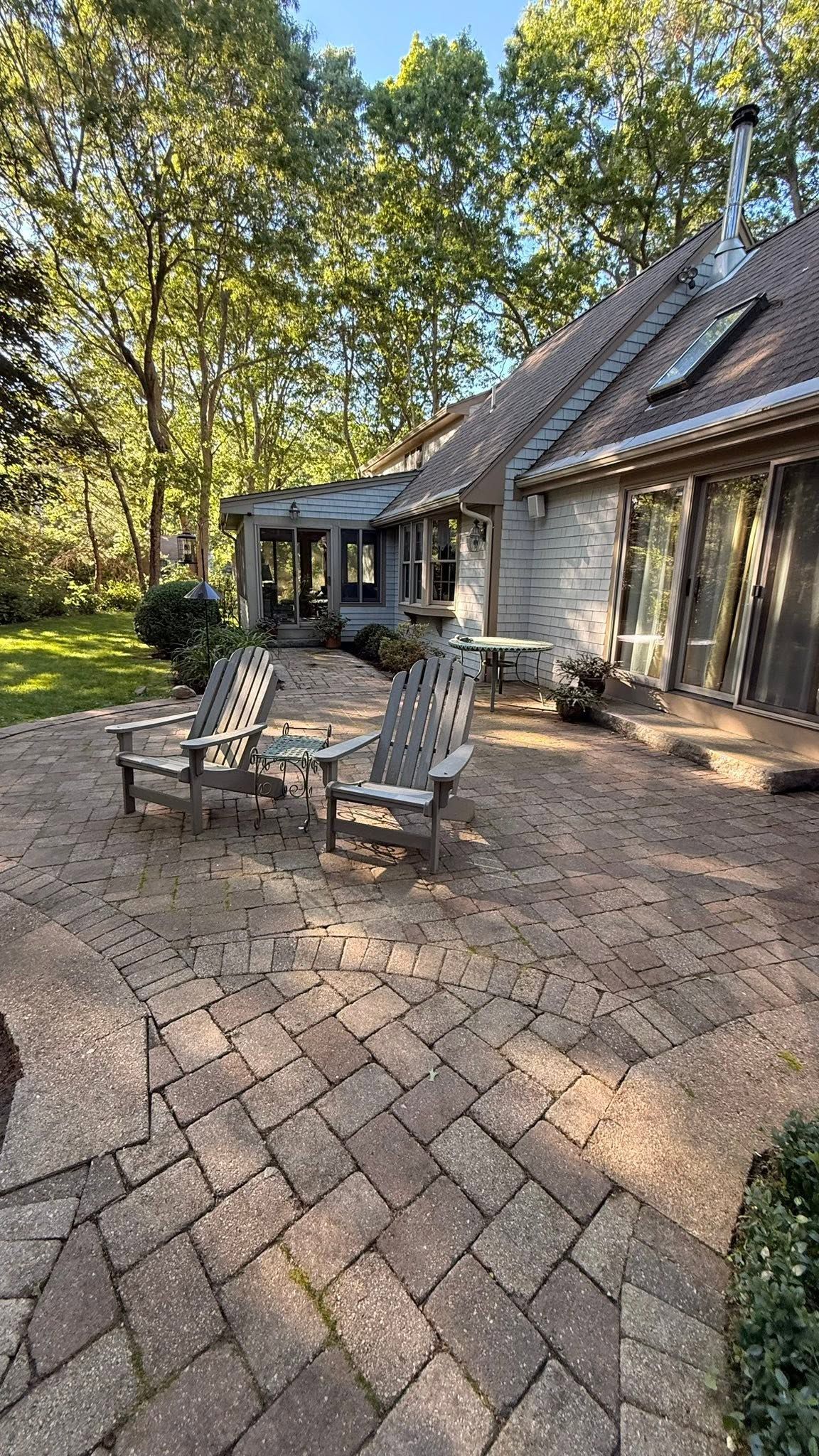 Brick patio with two chairs outside a stone house, surrounded by trees and greenery.