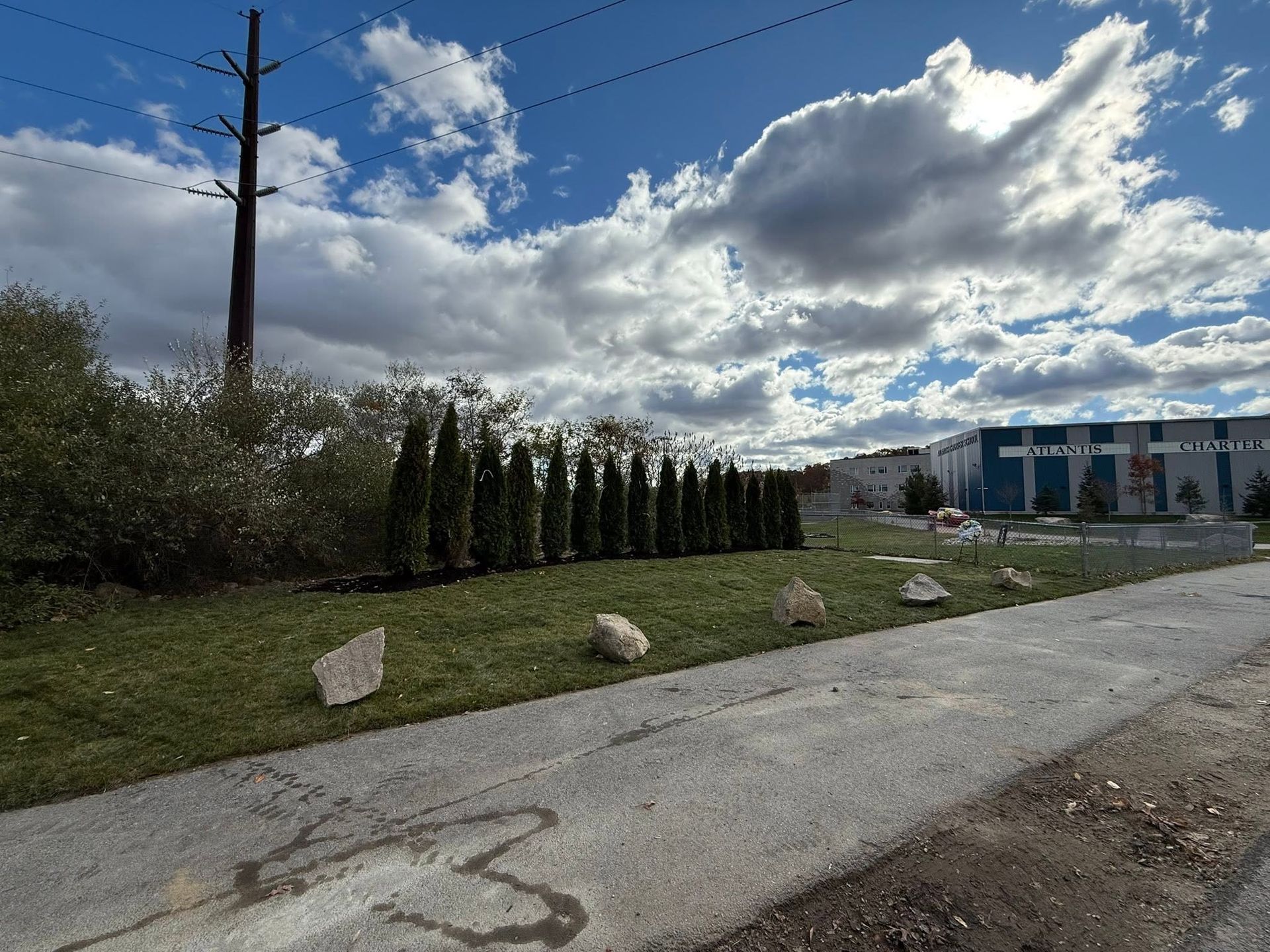 Rural roadside with a hedge, utility pole, cloudy sky, and a blue building in the distance.