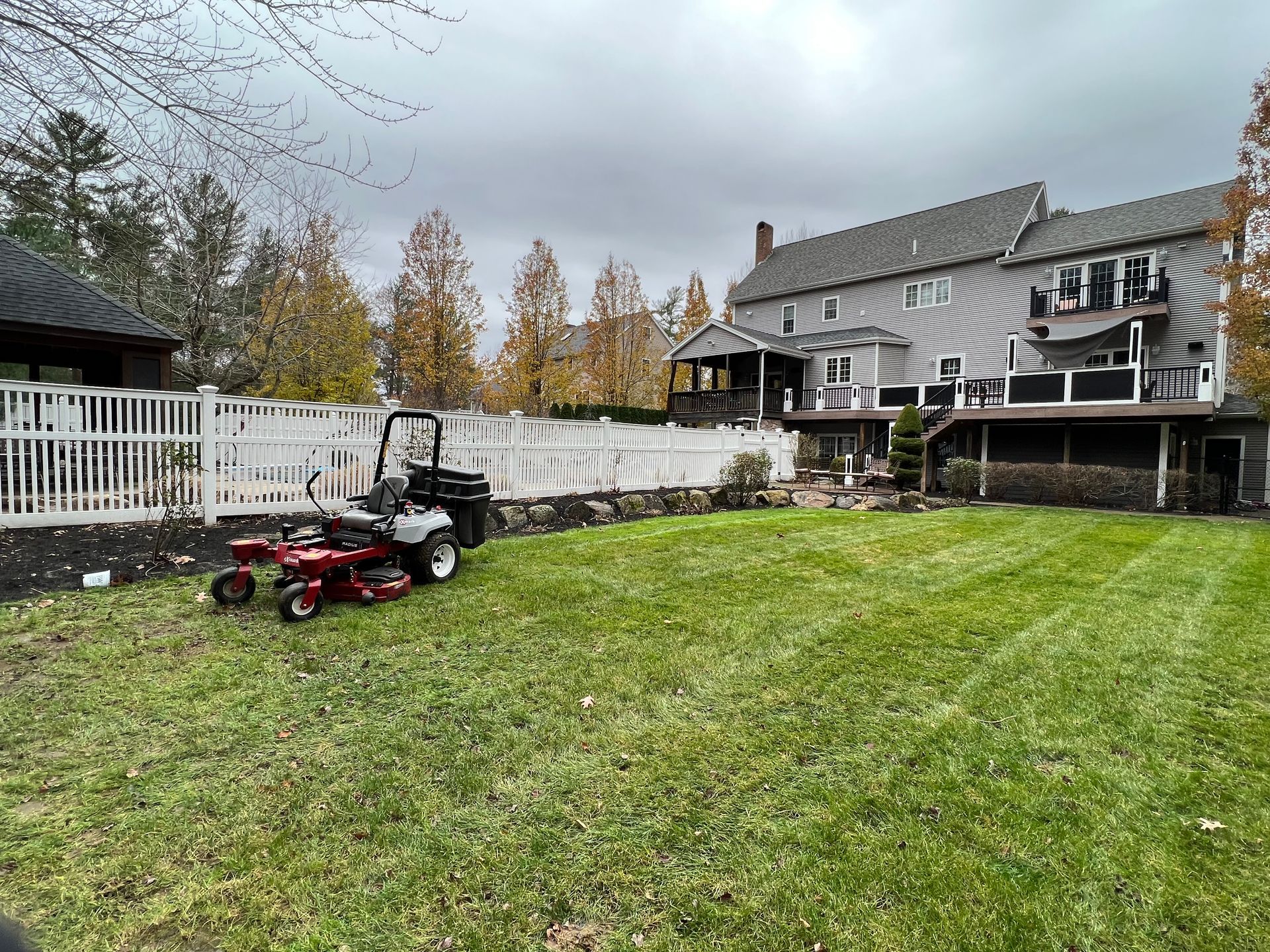 Backyard with green lawn, red lawn tractor, white fence, and two-story house in autumn.