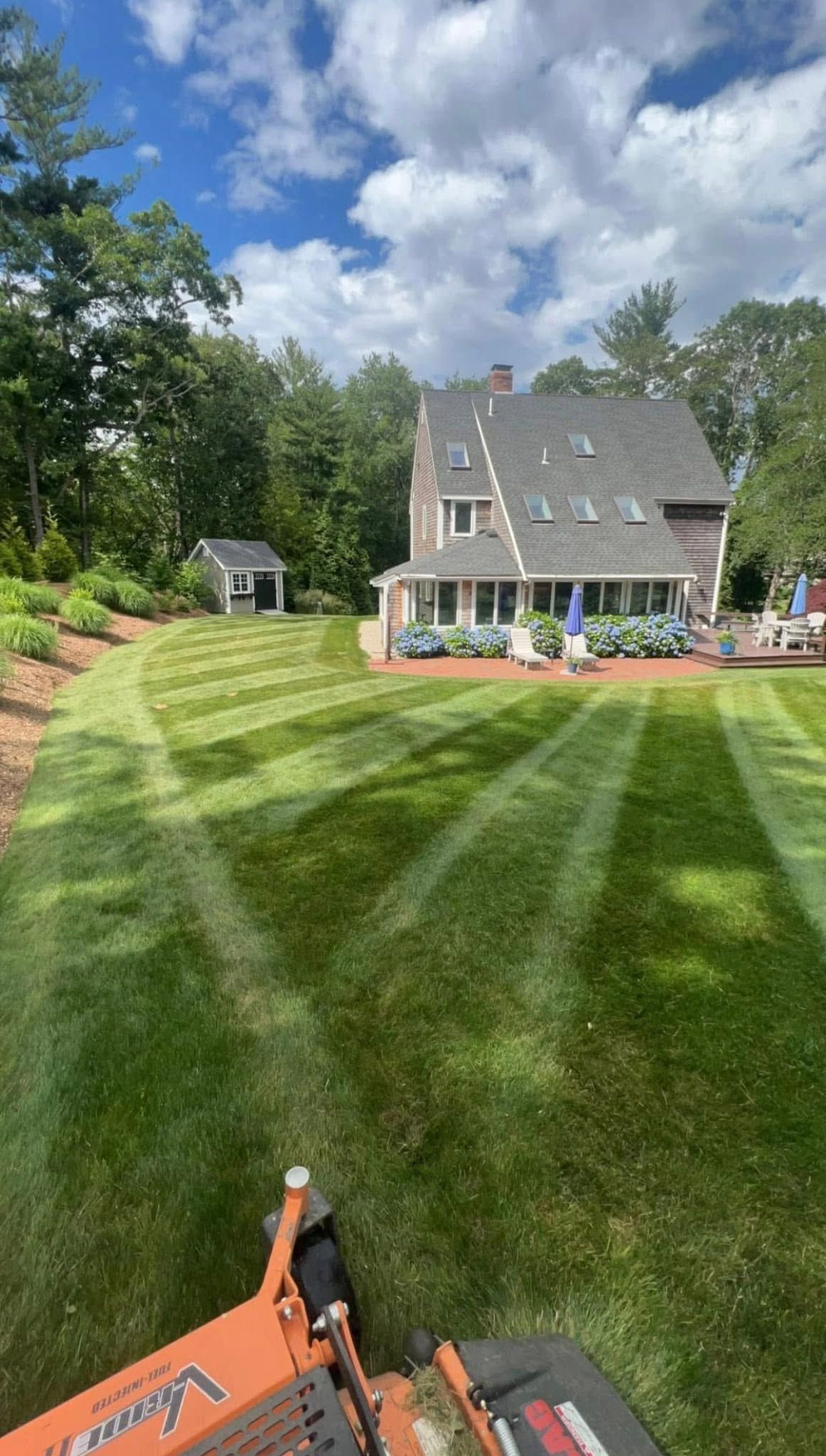 Freshly mowed grassy backyard with a small house, trees, and partly cloudy sky