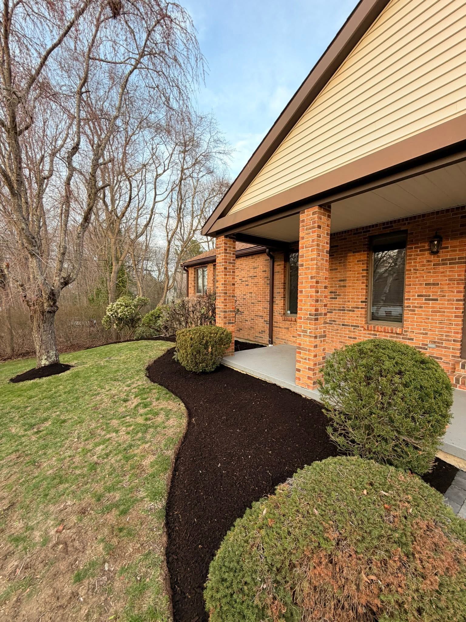 Brick house exterior with curved mulch beds, trimmed shrubs, and leafless trees along the lawn.
