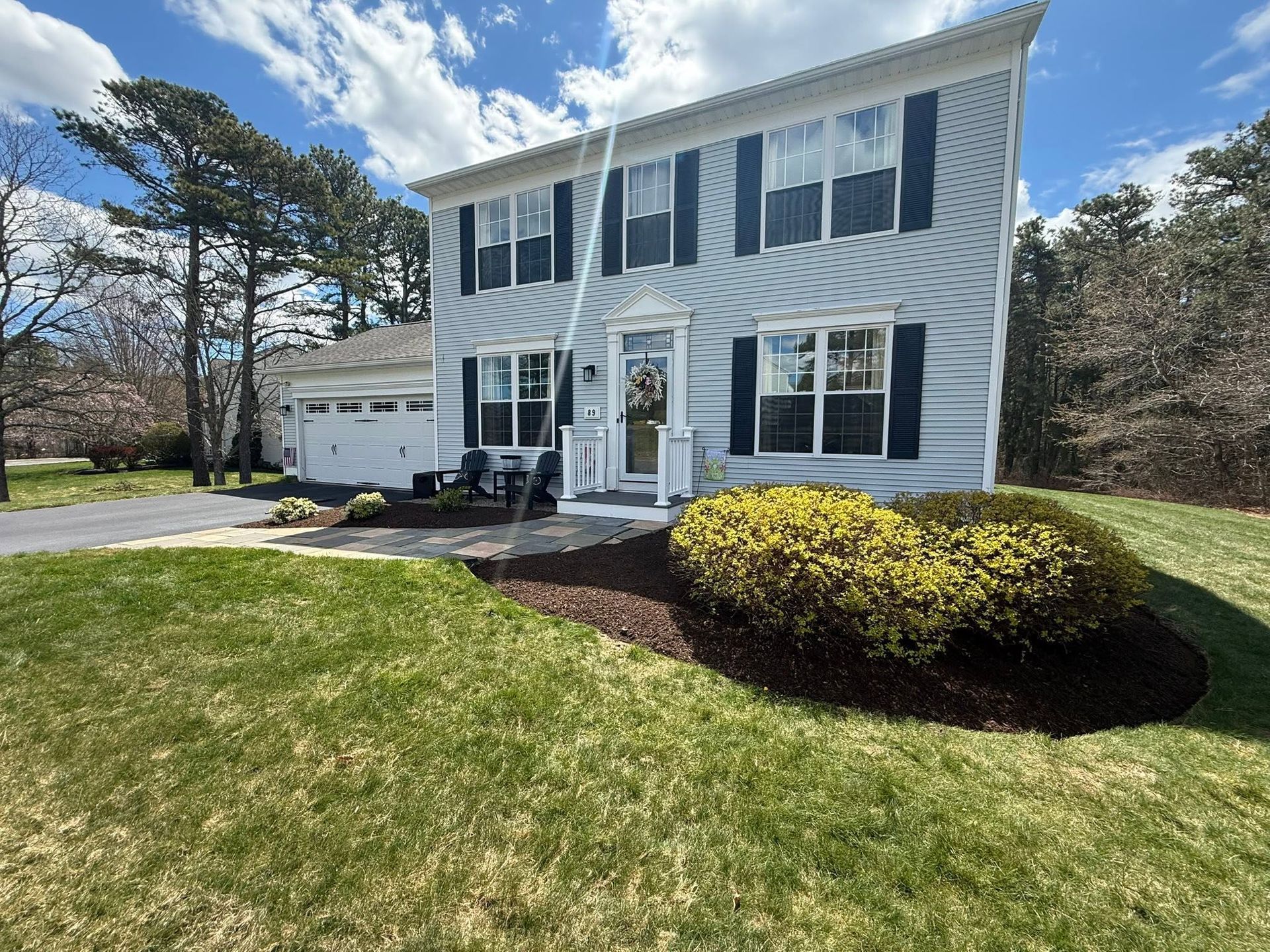 Two-story white house with blue shutters, garage, and landscaped front yard under a sunny sky