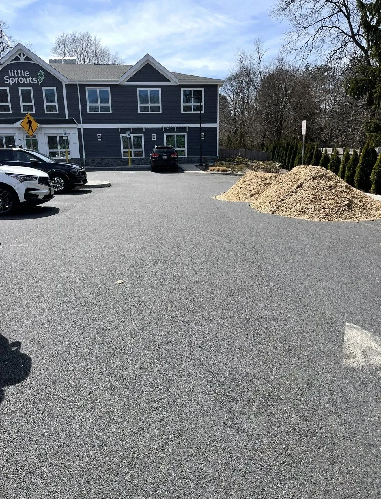 Parking lot beside a dark gray building, with parked cars and a large pile of wood chips on the right.