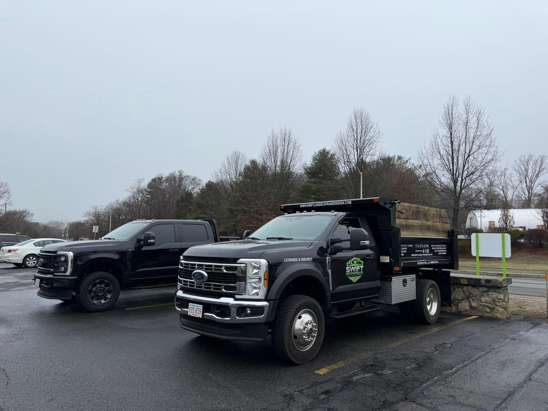 Black utility truck parked in a wet lot near another truck and a signboard on an overcast day